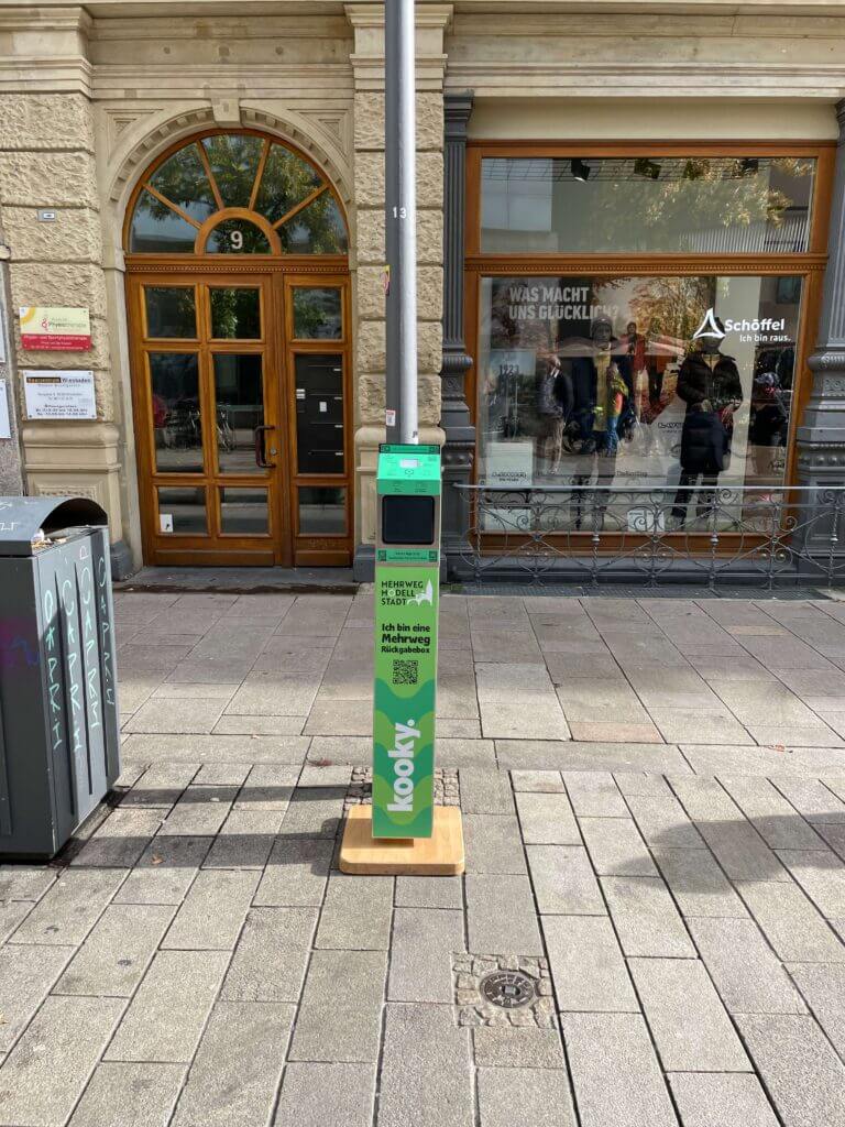 Eine grüne Rückgabebox vor einem Kleidungsgeschäft, die als Teil des Projekts Mehrweg Modell Stadt in Wiesbaden aufgestellt wurde.